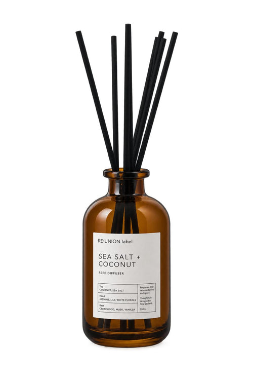 Amber glass reed diffuser bottle with a white label reading 'RE:UNION label SEA SALT + COCONUT' and eight black reed sticks, shown on a white background.