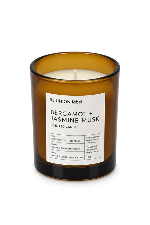 An amber glass jar candle with a white label reading 'RE:UNION label BERGAMOT + JASMINE MUSK SCENTED CANDLE' on a clean white background.