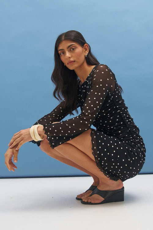 A woman crouches in a studio setting wearing a black long-sleeve mini dress with white polka dots and black wedge sandals against a blue backdrop.