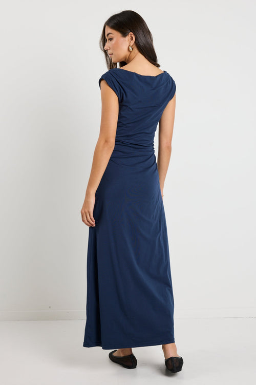 Woman wearing a navy maxi dress styled as a cowl neck and one-shoulder look, highlighting its versatile design and elegant ruched detailing.