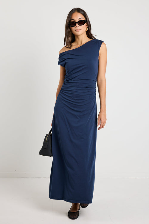 Woman wearing a navy maxi dress styled as a cowl neck and one-shoulder look, highlighting its versatile design and elegant ruched detailing.