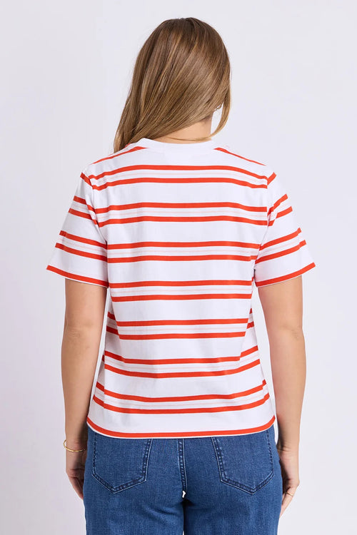 A woman viewed from the back wearing a white short-sleeve T-shirt with thin orange horizontal stripes and blue jeans, against a plain light gray studio background.