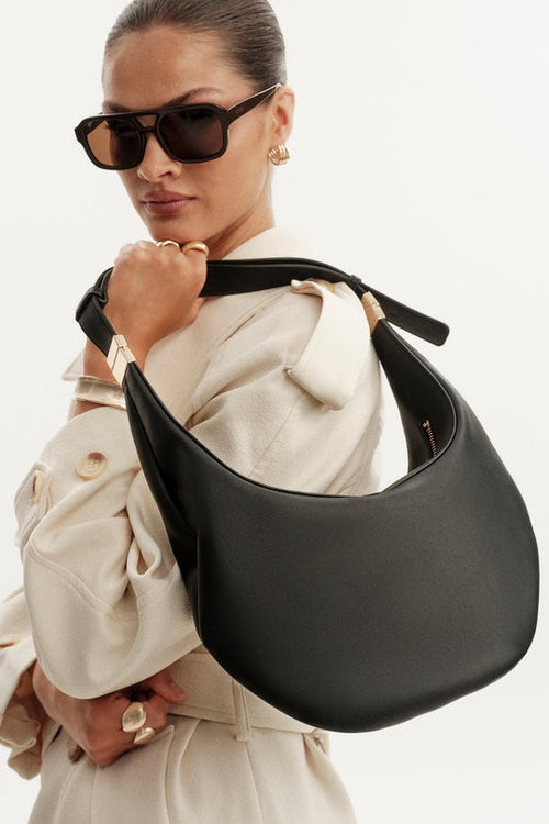 A model wearing a cream trench coat and black sunglasses carries a large, crescent-shaped black leather shoulder bag with gold-tone hardware against a plain white background.
