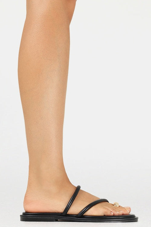 A side view of a person's foot and lower leg wearing a minimalist black flat slide sandal with a single strap and a small gold ring detail on the toe, set against a plain white background.