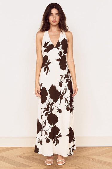 A woman modeling a sleeveless halter-neck maxi dress with a dark brown floral print on an off-white background, standing against a plain white wall on a light wood floor.