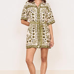A woman modeling a short-sleeve button-front mini dress with an olive green and cream botanical pattern, standing on a light wood herringbone floor against a plain off-white wall.