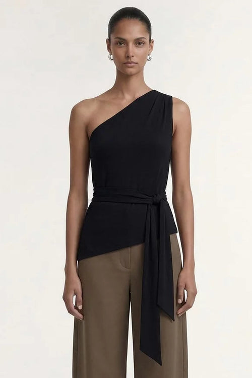 A model stands against a plain light background wearing a one-shoulder black wrap top and wide-leg taupe-brown pants with black sandals.