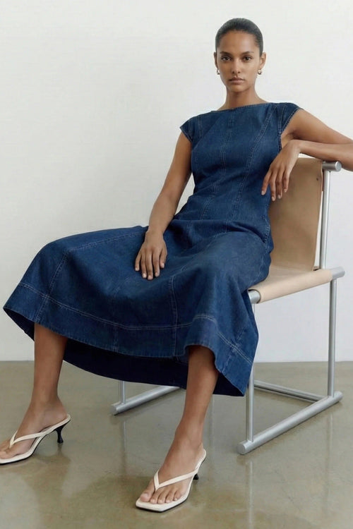 A woman wearing a dark blue denim midi dress sits on a minimalist beige and silver chair in a studio setting.