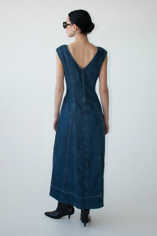 A woman viewed from the back wearing a dark blue denim maxi dress with a deep V-neckline and a buttoned back, paired with black pointed-toe heels against a plain white studio background.