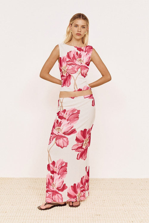 A woman modeling a sleeveless white maxi dress with a bold pink floral print, standing against a clean off-white studio background.
