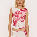 A woman modeling a sleeveless white dress with a bold pink floral print, featuring a high neckline, a midriff cutout, and a matching skirt, set against a plain light background.