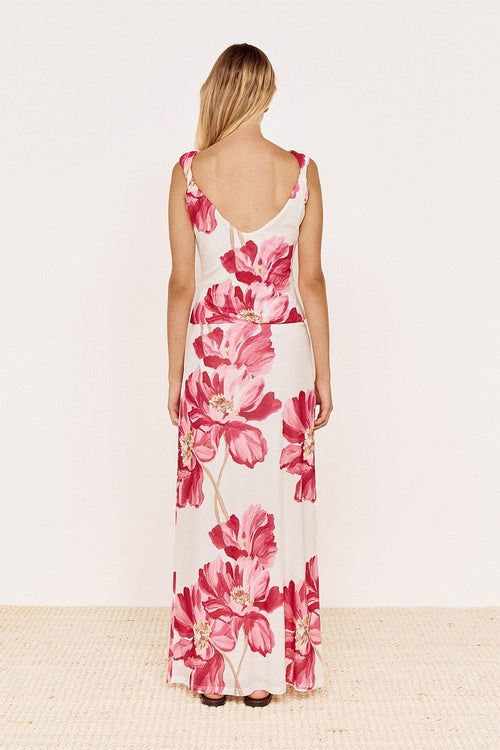 A woman viewed from the back wearing a sleeveless maxi dress with a deep V-neckline and a bold pink floral print on a white base, standing against a plain off-white wall.