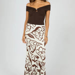 A woman modeling a two-piece outfit consisting of a dark brown off-the-shoulder top with a small front keyhole detail and a matching long skirt with a brown abstract floral pattern on an off-white background, set against a light gray studio backdrop.