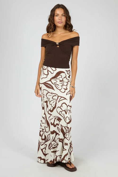 A woman modeling a two-piece outfit consisting of a dark brown off-the-shoulder top with a small front keyhole detail and a matching long skirt with a brown abstract floral pattern on an off-white background, set against a light gray studio backdrop.