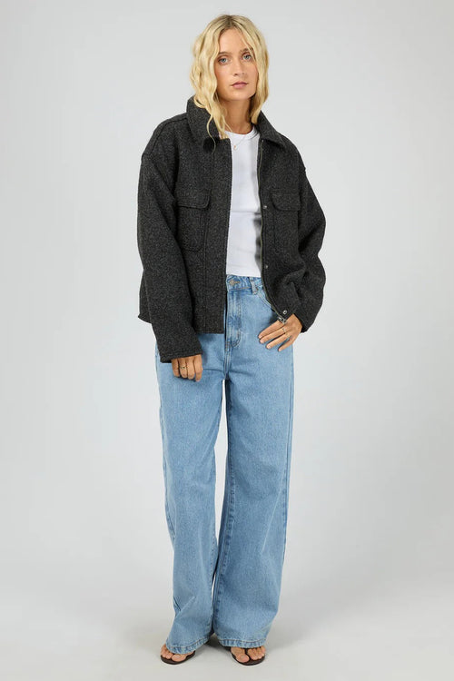 Woman wearing a dark jacket, white shirt, and light blue jeans on a plain background