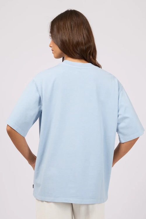 A woman shown from the back wearing an oversized light blue t-shirt and cream-colored pants, photographed against a solid light gray studio background.