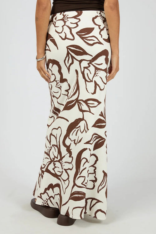 A person wearing a long skirt with a brown floral and leaf pattern on an off-white background, shown from the back against a light gray studio backdrop.