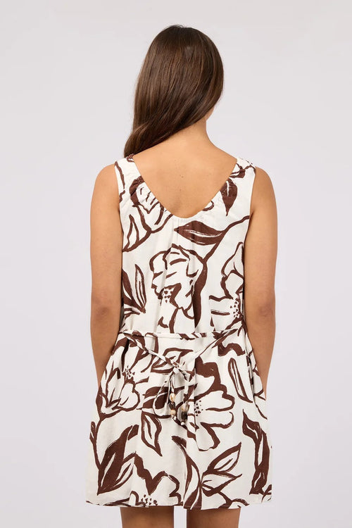 A woman viewed from behind wearing a sleeveless cream dress with a brown floral and leaf pattern, tied at the waist, against a light gray studio background.