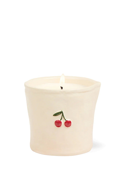 An off-white ceramic candle holder with a small cherry decoration on the front, containing white wax and a lit flame, set against a plain white background.