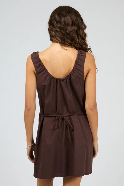 A woman shown from the back wearing a sleeveless brown dress with a gathered open back and a tie detail at the waist, photographed against a light gray studio background.