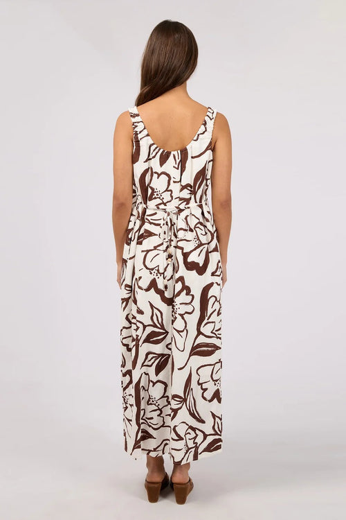 A woman viewed from the back wearing a sleeveless maxi dress with a brown floral line-art print on a cream background, paired with brown heeled sandals, against a light gray studio backdrop.