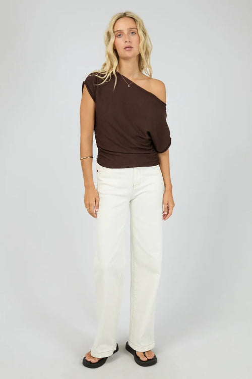 A woman modeling a dark brown off-the-shoulder knit top paired with white wide-leg pants and black sandals, standing against a light gray studio background.