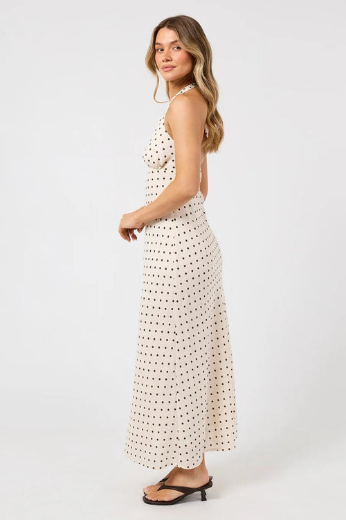 A woman modeling a sleeveless cream maxi dress with a small black polka dot pattern, shown from a side-back angle against a plain white studio background.