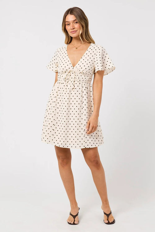 A woman modeling a short-sleeve cream dress with a black polka dot pattern, featuring a V-neckline, ruffled sleeves, and a tie detail at the waist, set against a plain light gray studio background.