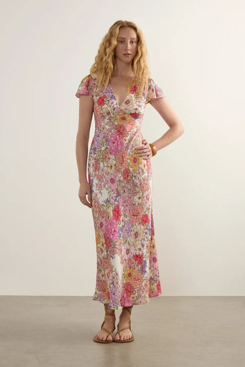 A woman modeling a short-sleeve, V-neck maxi dress with a dense pink, red, yellow, and blue floral pattern on a light cream background, set against a neutral studio backdrop.