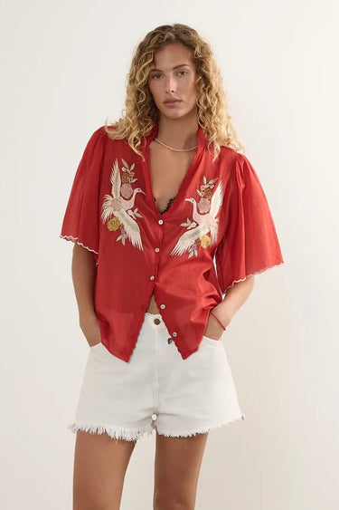 A woman stands in a studio wearing a red short-sleeve button-up shirt with white and gold butterfly embroidery, paired with white denim shorts and white slide sandals.