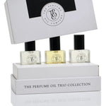Three small perfume oil bottles displayed in a white presentation box with the lid propped behind, on a clean white background. The packaging reads 'THE PERFUME OIL TRIO COLLECTION' and shows a circular monogram logo.