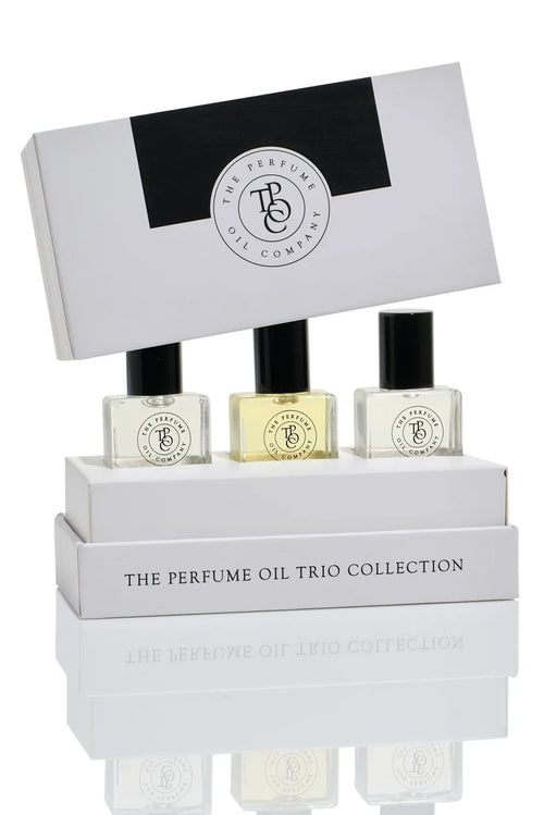 Three small perfume oil bottles displayed in a white presentation box with the lid propped behind, on a clean white background. The packaging reads 'THE PERFUME OIL TRIO COLLECTION' and shows a circular monogram logo.