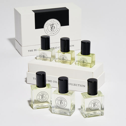 Three small perfume oil bottles with black caps arranged in front of white branded boxes on a clean white studio background. The bottles contain pale yellow liquid, and the packaging displays the brand name 'The Perfume Oil Collection' with a circular 'BC' logo.