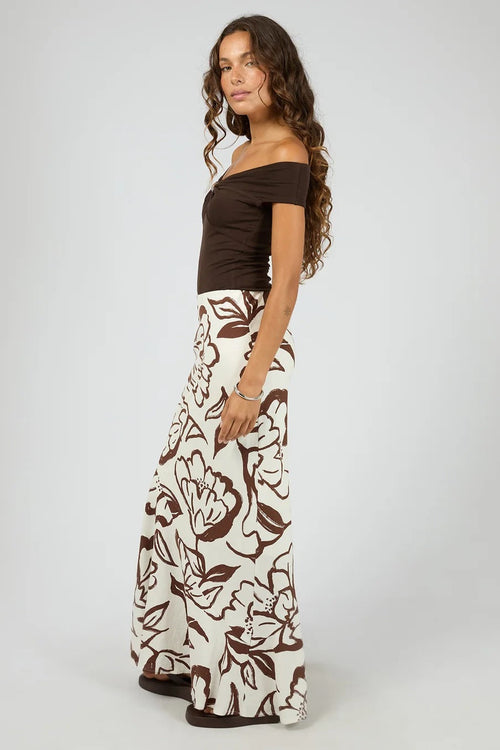 A woman modeling a two-piece outfit consisting of a dark brown off-the-shoulder short-sleeve top and matching dark brown floral print wide-leg pants on a light gray studio background.