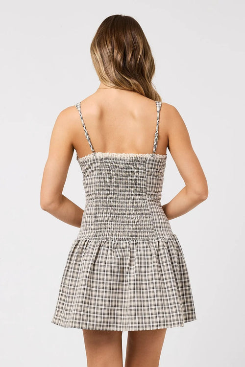 A woman shown from the back wearing a sleeveless mini dress with thin straps, a smocked bodice, and a flared skirt in a beige and gray plaid pattern against a light gray studio background.