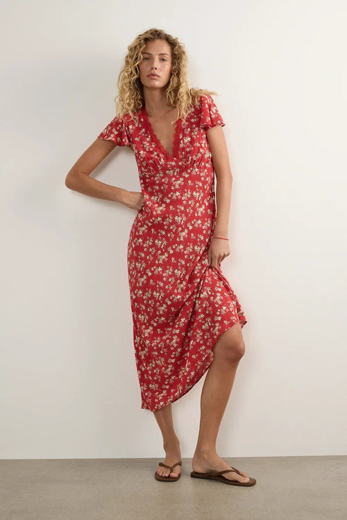 A woman modeling a red short-sleeve wrap midi dress with a white floral pattern, standing against a plain off-white wall.
