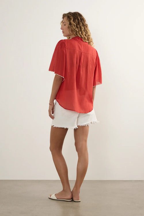 A woman shown from the back wearing a loose coral-red short-sleeve blouse and white frayed denim shorts, standing against a plain light gray studio wall.