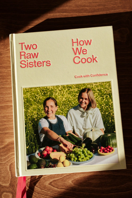 The book 'Two Raw Sisters: How We Cook' lies on a dark wooden surface under warm, direct sunlight. The cover is cream-colored with bold red text and a central photograph of two women surrounded by fresh vegetables.