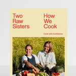 The front cover of the cookbook 'Two Raw Sisters: How We Cook' is shown standing upright against a clean white background. The cover features a pale yellow background with red text and a central photograph of two women outdoors in a field of yellow flowers, surrounded by fresh produce.