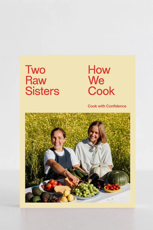 The front cover of the cookbook 'Two Raw Sisters: How We Cook' is shown standing upright against a clean white background. The cover features a pale yellow background with red text and a central photograph of two women outdoors in a field of yellow flowers, surrounded by fresh produce.