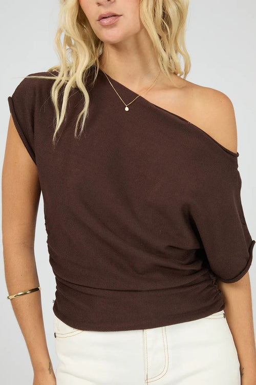 A woman wearing a dark brown off-the-shoulder knit top with elbow-length sleeves, paired with white pants, against a light gray studio background.
