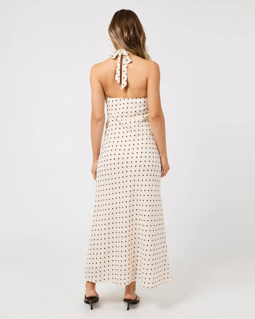 A woman viewed from the back wearing a halter-neck maxi dress with a small black polka dot pattern on a warm off-white background, paired with black pointed-toe heels, against a plain white studio background.