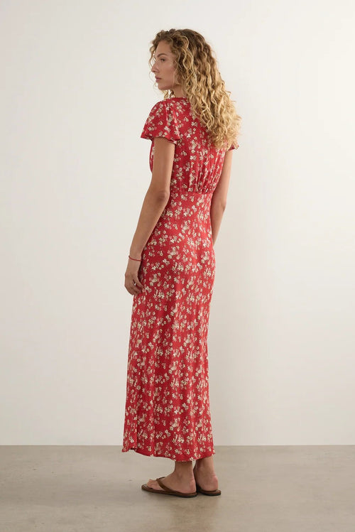 A woman with curly blonde hair is shown from the back wearing a red maxi dress with a white floral pattern, standing against a plain off-white wall.