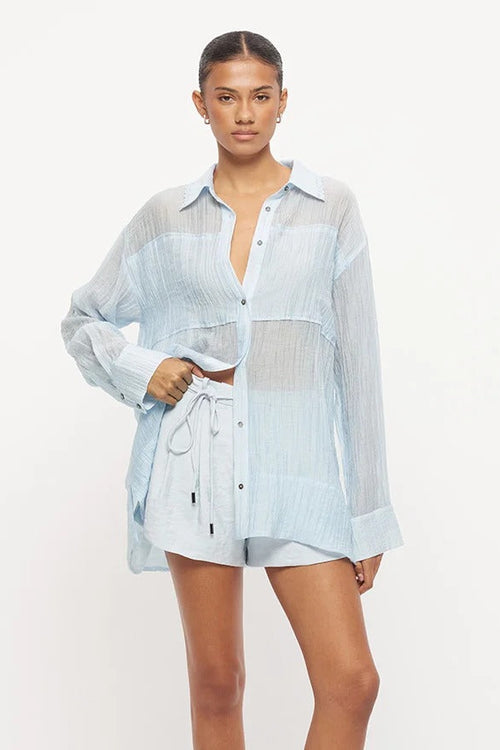 Model wearing a light blue semi-sheer oversized button-up shirt with long sleeves, paired with matching drawstring shorts and flat sandals.