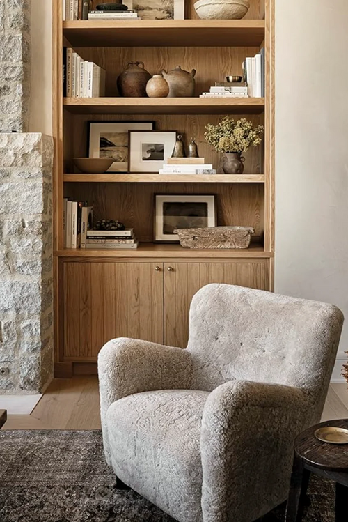 A cozy living room corner featuring a light beige bouclé armchair in the foreground, with a tall built-in wooden bookshelf behind it styled with books, vases, and framed art.