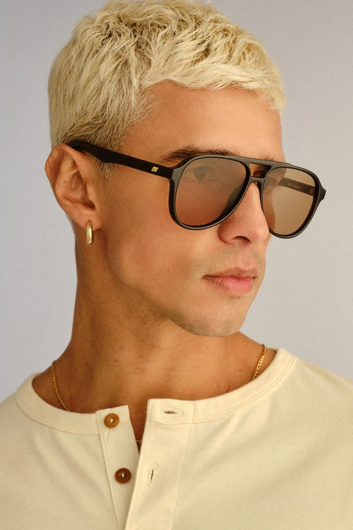 A man with short blonde hair wearing black aviator-style sunglasses with brown lenses and a cream henley shirt, shown in a close-up profile against a light gray background.