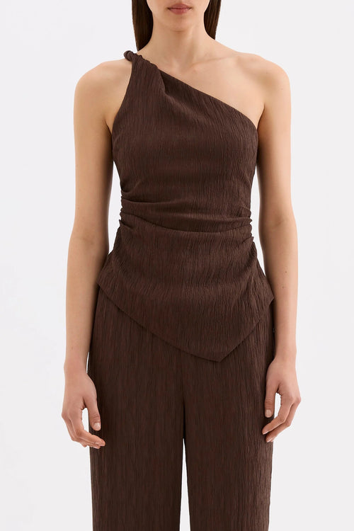 A model wearing a dark brown crinkled one-shoulder jumpsuit with a draped waist and matching wide-leg pants, shot against a plain white studio background.
