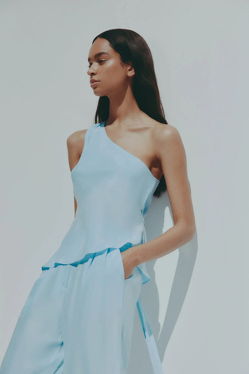 A woman modeling a light blue one-shoulder jumpsuit with a matching fabric belt, set against a plain light gray background.
