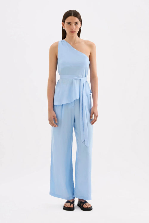 A woman modeling a light blue one-shoulder wrap jumpsuit with wide-leg pants and black sandals, standing against a plain white studio background.