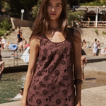 A woman wearing a sleeveless, dark brown crochet lace dress stands on a sunny beachside walkway, holding a striped knit bag.